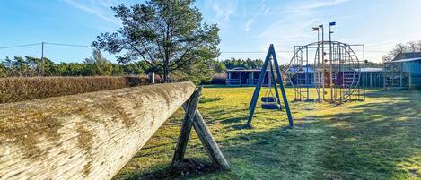Un parque infantil con columpios y una estructura para escalar, rodeado de un campo cubierto de césped y una valla de madera.