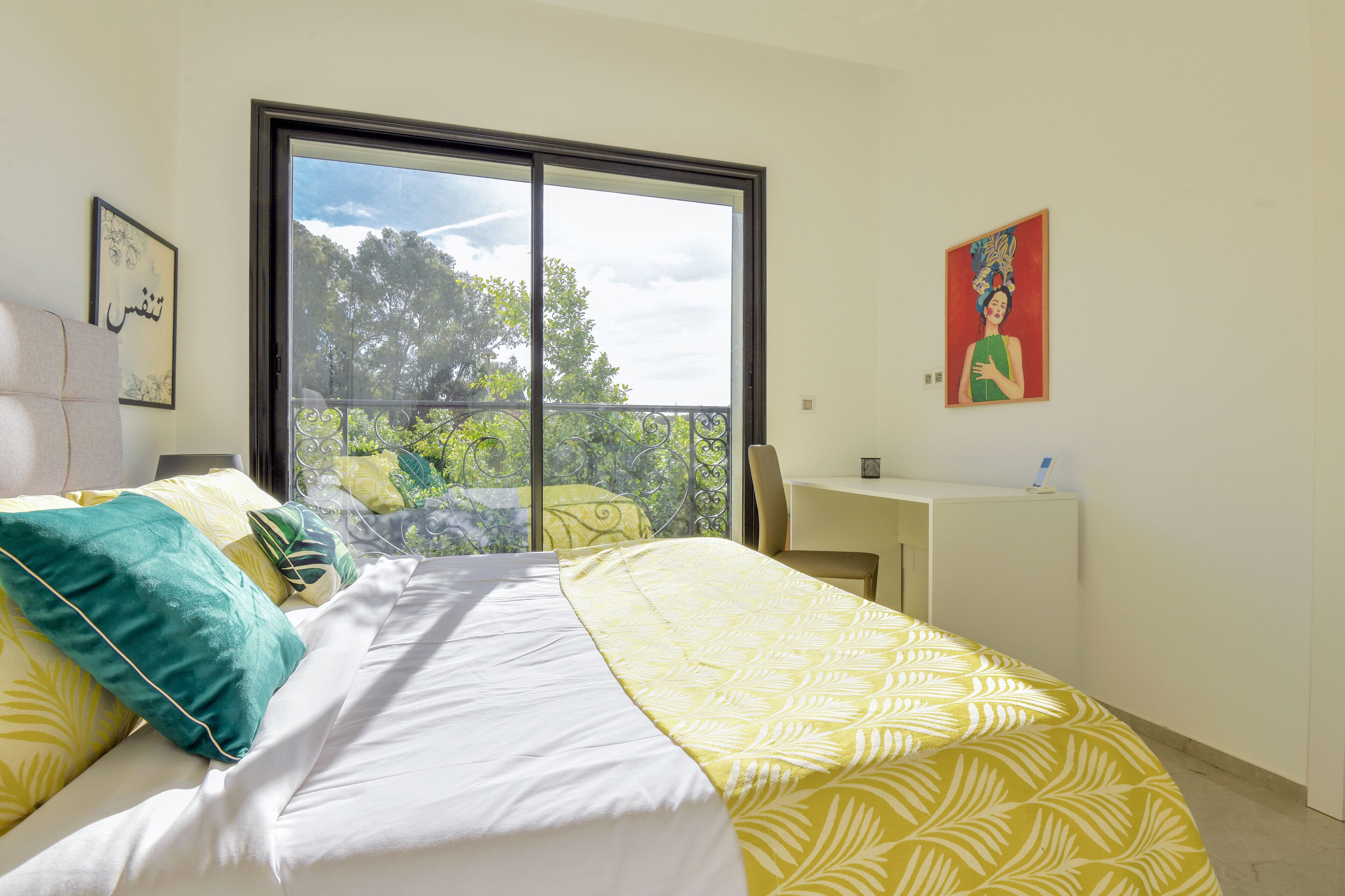 A bedroom with a bed, a desk, and a balcony with a view of trees.