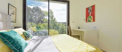 A bedroom with a bed, a desk, and a balcony with a view of trees.