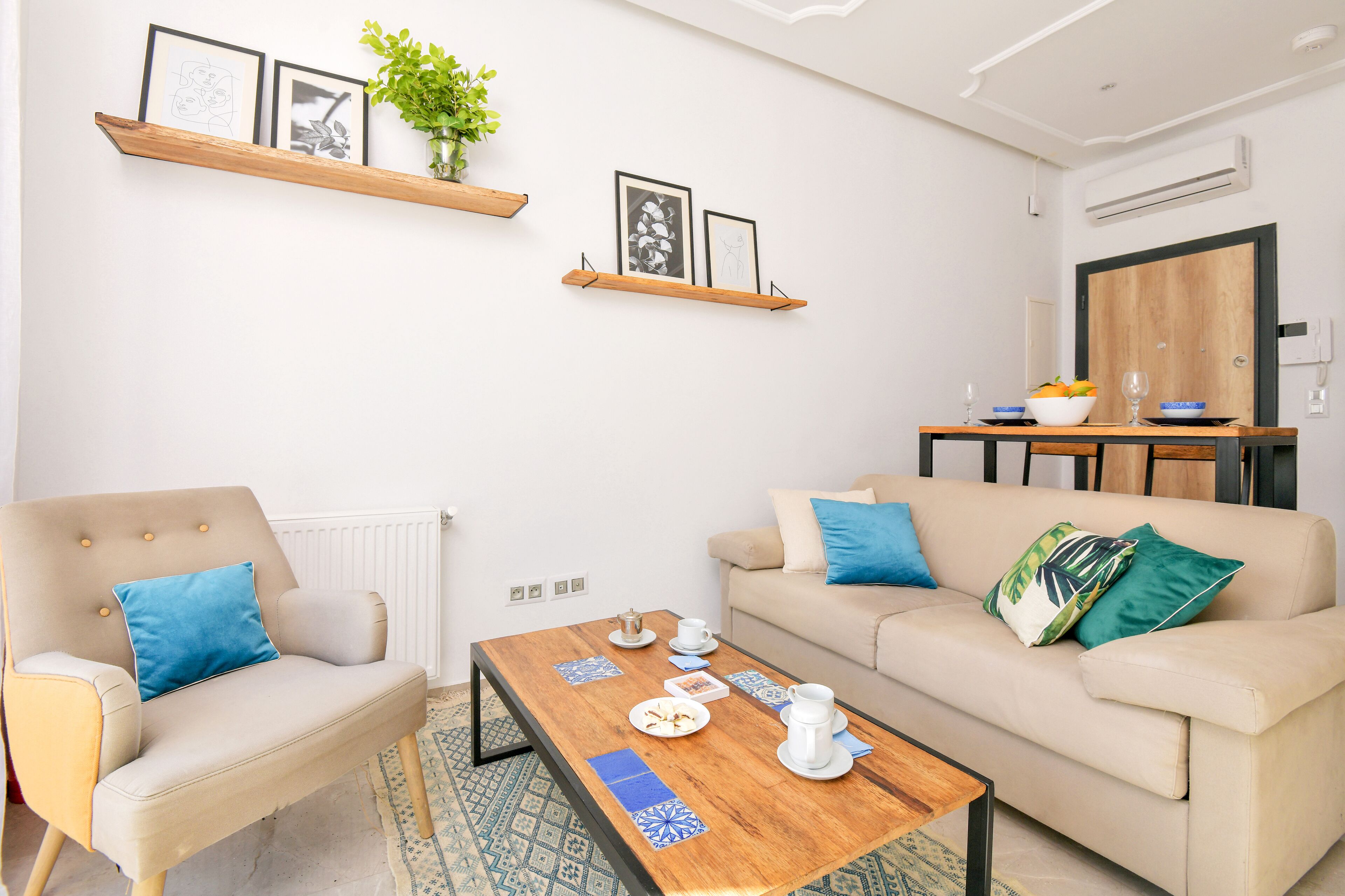 A living room with a sofa, armchair, coffee table, and a dining table with a bowl of fruit and a wine glass.