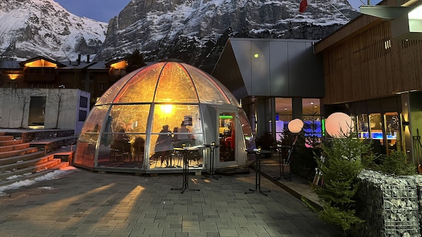 A glass dome structure with a view of a mountain range, a wooden building with a sloped roof, and flags in the background. - Great Views, Great Location, modern & cozy Apartment (renovated 2022) (Grindelwald)
