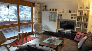 A modern living room with a large window, a sofa, a coffee table, a TV, and a bookshelf. - Great Views, Great Location, modern & cozy Apartment (renovated 2022) (Grindelwald)