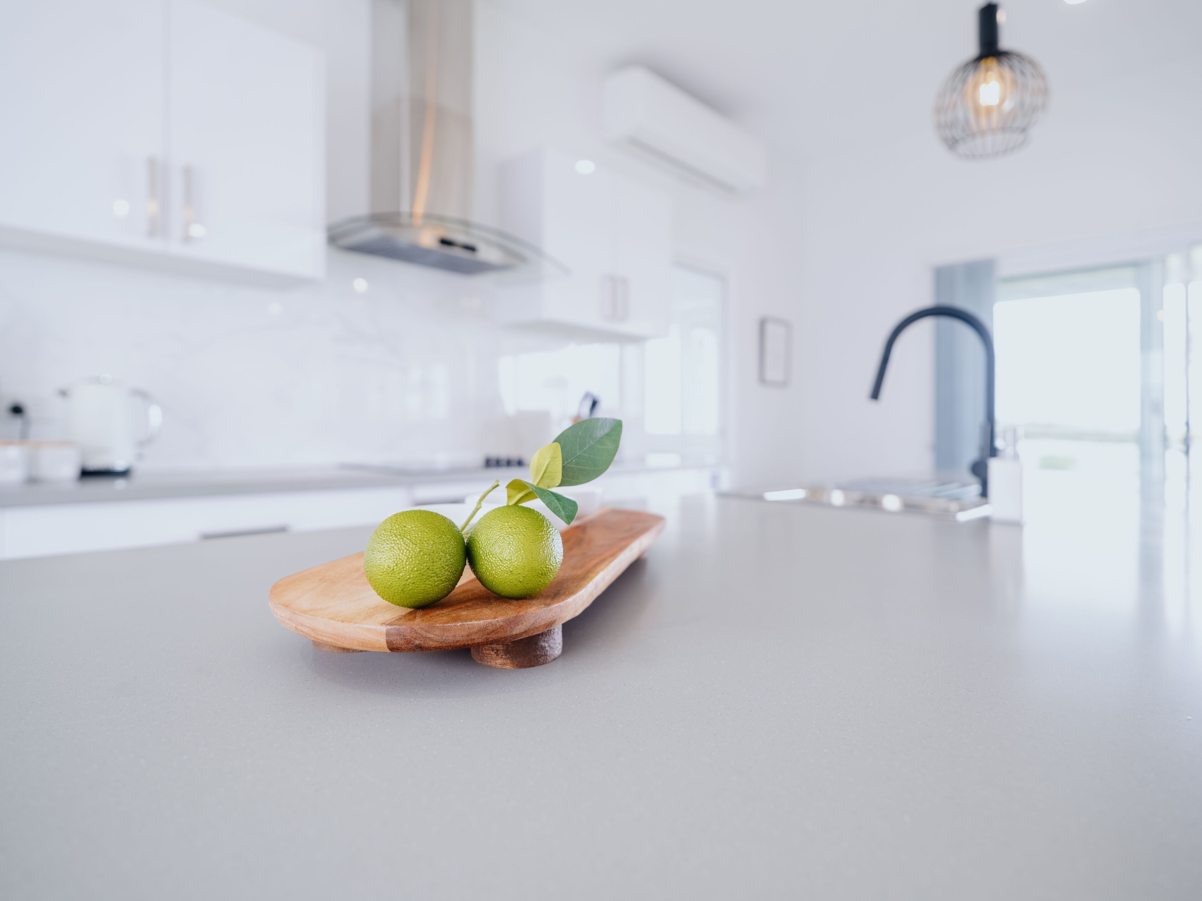 Cocina moderna con una tabla de madera que sostiene tres frutas verdes y una hoja, una campana extractora de acero inoxidable y una lámpara colgante.