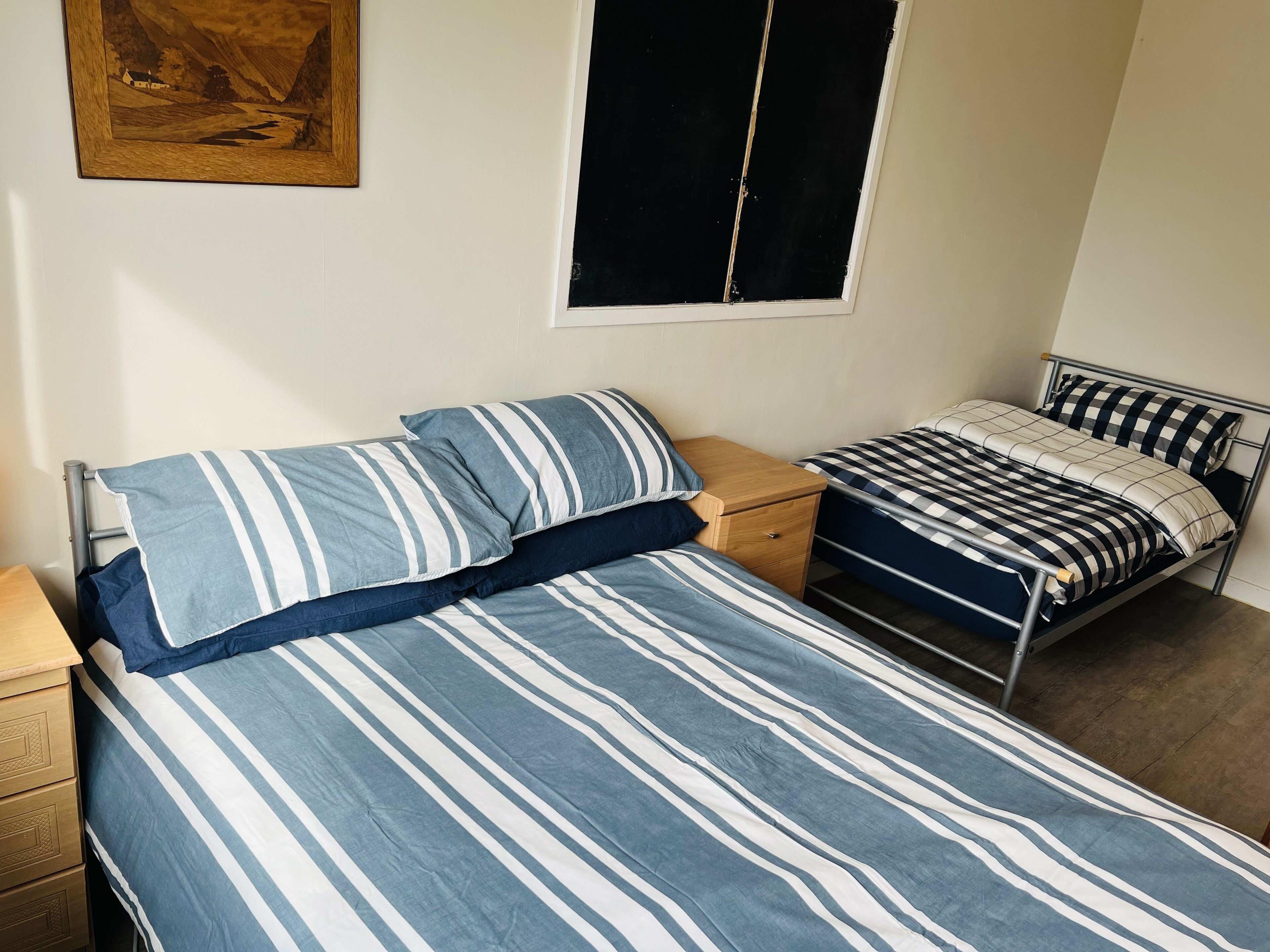 A bedroom with two beds, one larger and one smaller, both with striped bedding. There is a wooden nightstand with a drawer and a small table beside the larger bed. A framed picture hangs on the wall above the beds.