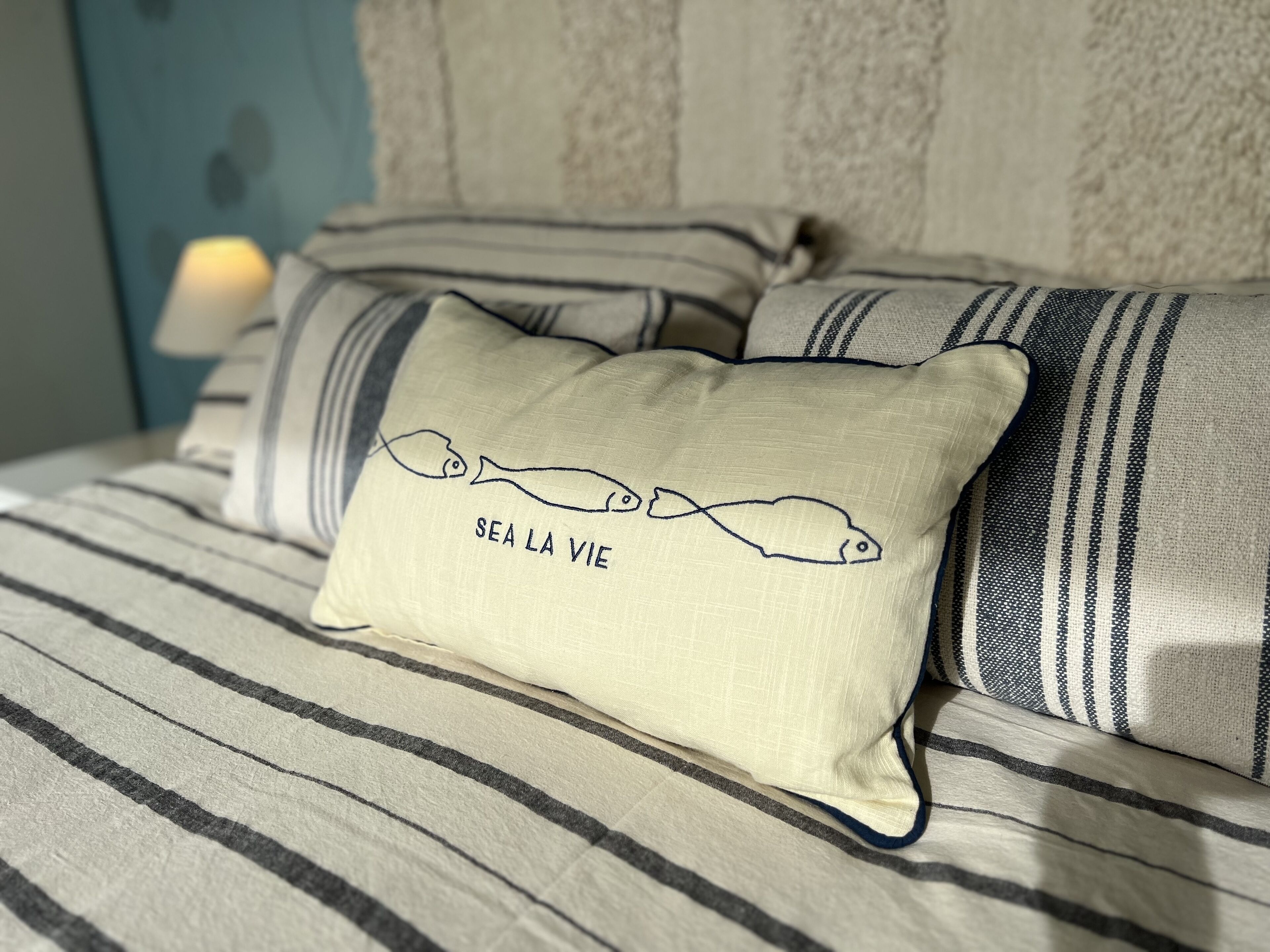 Uma cama com roupa de cama listrada e um travesseiro com ilustração de peixe e o texto "SEA LA VIE".