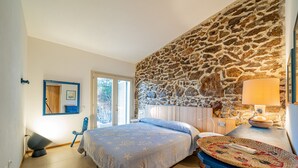 A bedroom with a stone wall, a bed with a blue bedspread, a bedside table with a lamp, and a view of the outdoors through a window. - Casa Loris by Wonderful Italy (Olbia)