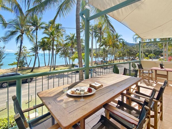 A wooden table with a plate of food and glasses, a green railing, and a view of the ocean and palm trees. - Sails on Horseshoe Apartment 4 (Horseshoe Bay)