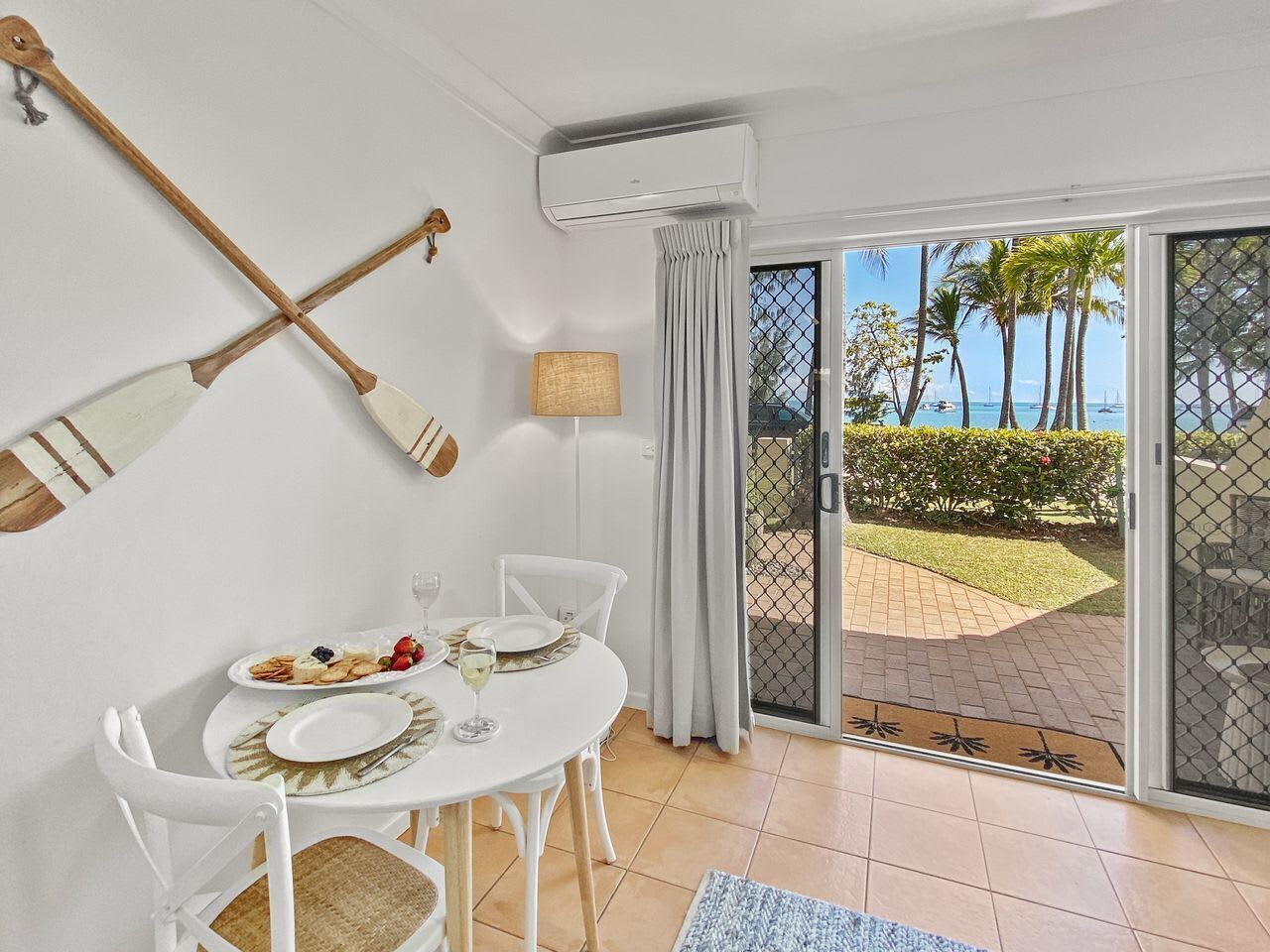 Un coin repas avec une table dressée pour deux personnes, une chaise blanche, une petite table avec une lampe, et une vue sur l’extérieur grâce à une porte-fenêtre coulissante.