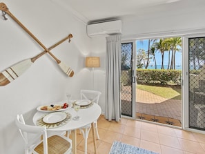 A dining area with a table set for two, a white chair, a small table with a lamp, and a view of the outdoors through a sliding glass door.
