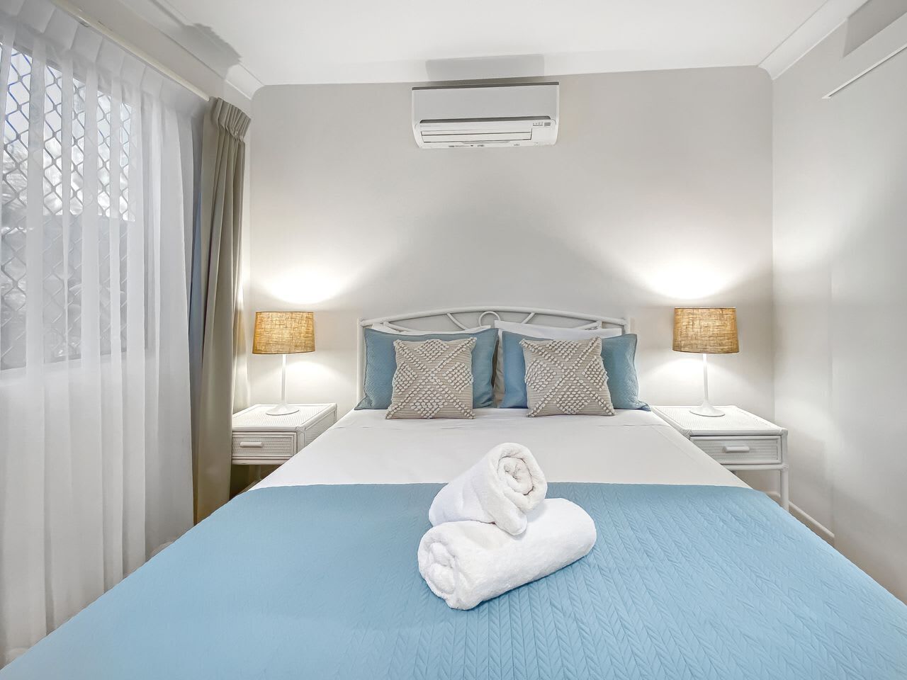 A hotel room with a bed, two bedside tables, a wall-mounted air conditioner, and a window with curtains.