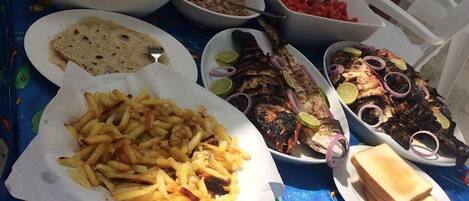 A table set with various dishes including grilled fish, fries, and bread, with a blue tablecloth featuring fish patterns.