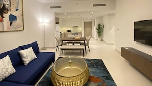 Un salon moderne avec un canapé bleu, une table basse en verre, un coin repas comprenant une table et des chaises, et un téléviseur fixé au mur.