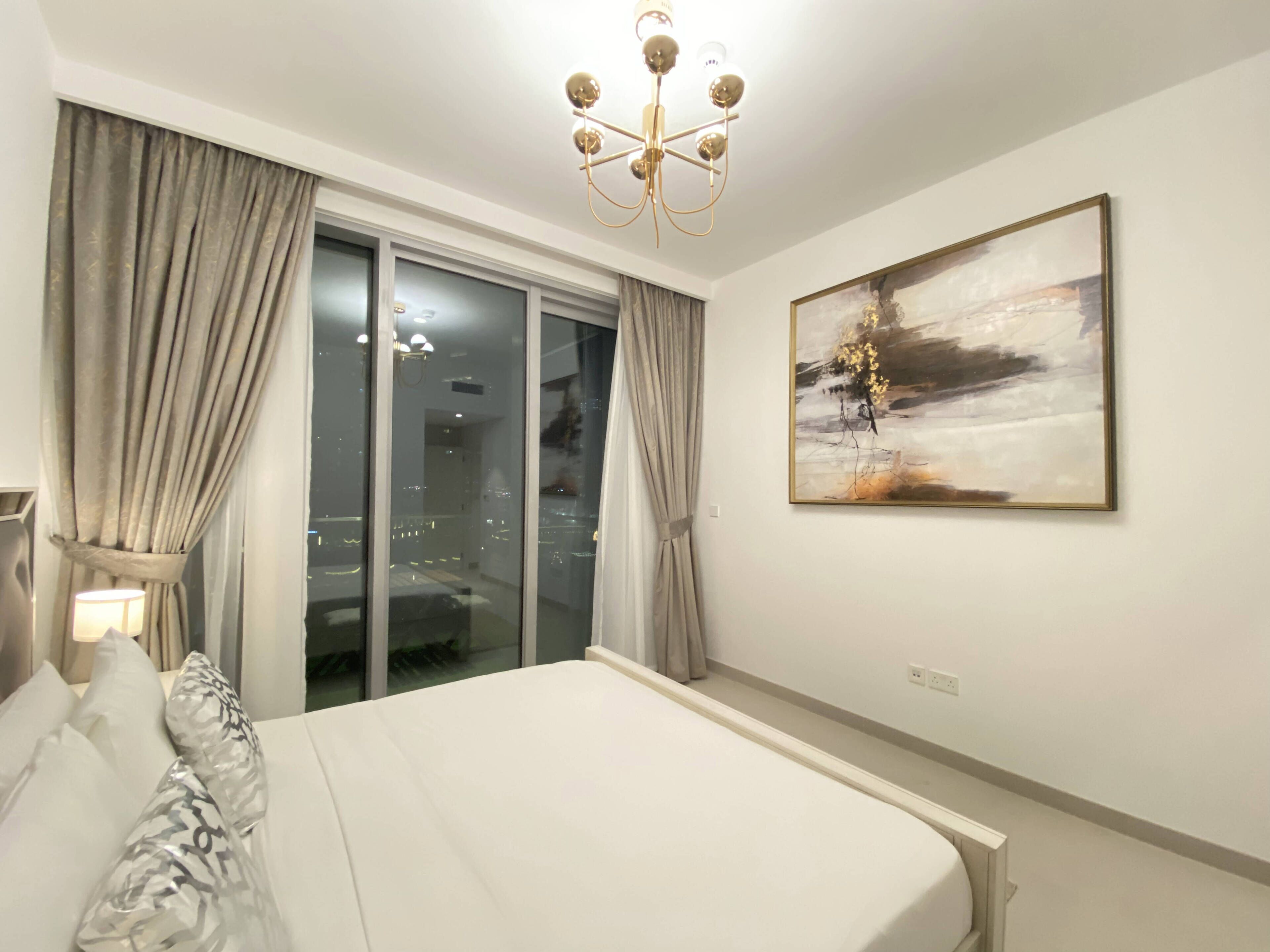 Une chambre à coucher avec un grand lit, un lustre, une porte-fenêtre coulissante et un tableau au mur.