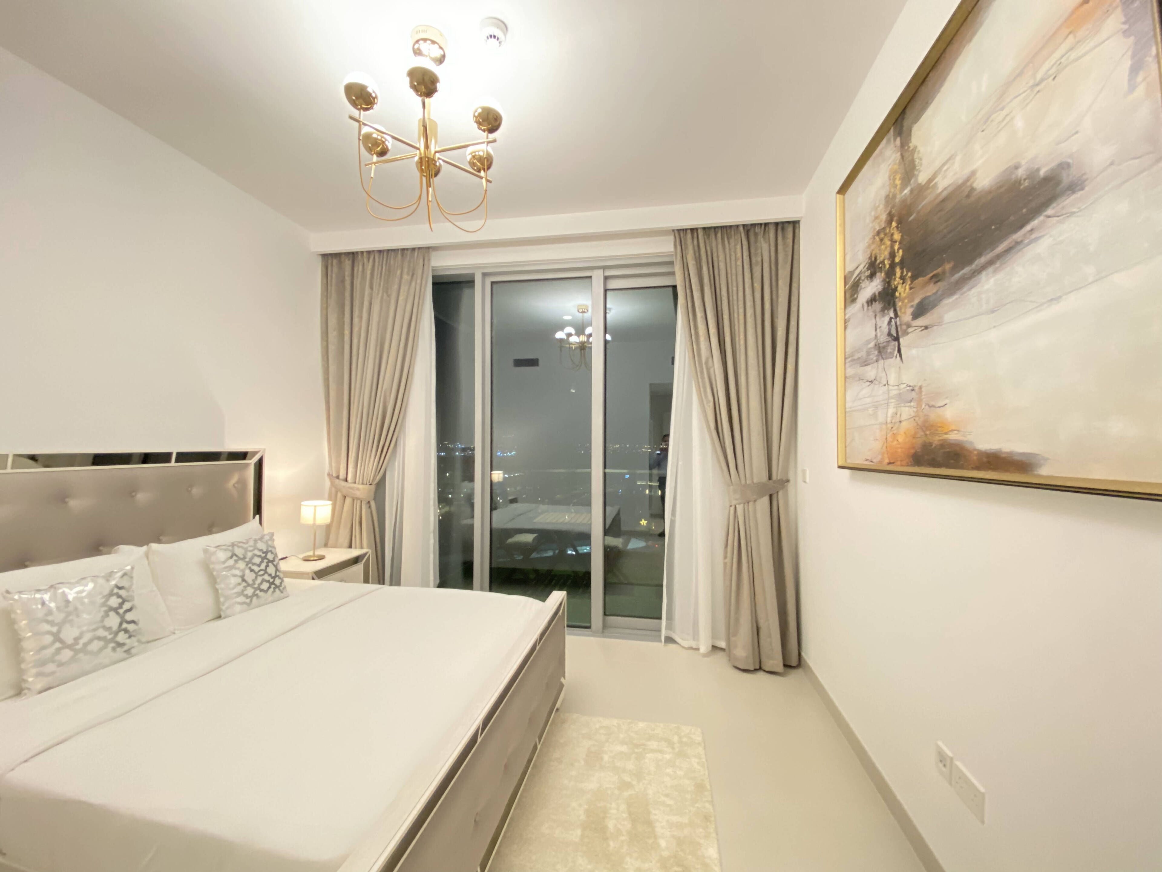 A bedroom with a large bed, a chandelier, a painting, and a view of the city through the window.