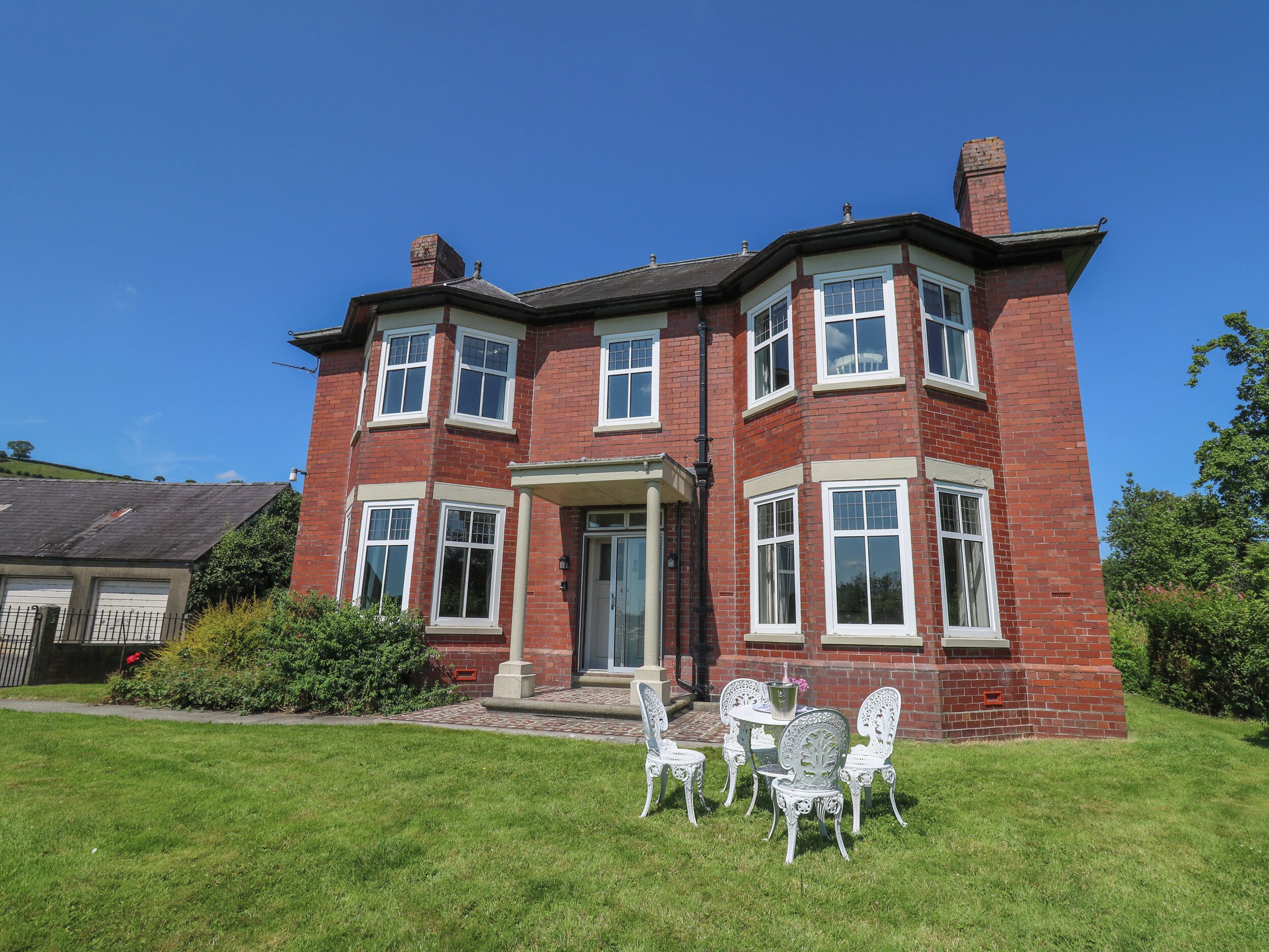 A two-story red brick house with a front porch, white-framed windows, and a small garden with white chairs.