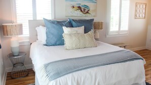 A neatly made bed with a white and blue bedspread, two side tables with lamps, and a painting of boats on the wall.