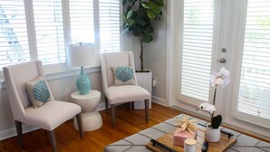 A room with two white armchairs, a small round table, a wooden coffee table with a tray, a potted plant, and a window with blinds.
