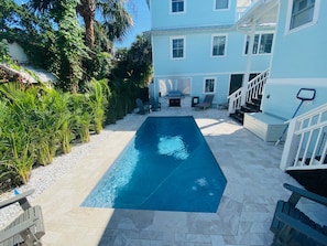 A rectangular swimming pool with a stone deck, surrounded by a white house with multiple windows and a covered outdoor area with chairs and a grill.