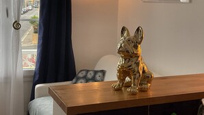 Une pièce avec une table en bois, une statuette de chien décorative, une fenêtre avec des rideaux et un luminaire fixé au mur.