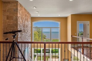 Una habitación con un telescopio sobre un trípode, un amplio ventanal de arco con vista al mar y una escalera con barandilla.