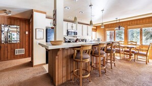 Een keuken met houten kastjes, een centraal eiland met krukken, hanglampen en uitzicht op het buitenleven door grote ramen.