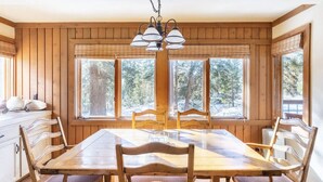 Een houten eetkamer met een grote tafel en stoelen, een hanglamp en grote ramen met uitzicht op bomen.
