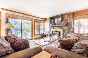 A cozy living room with a stone fireplace, comfortable seating, and large windows.