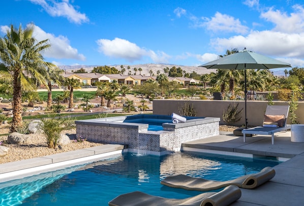 A swimming pool with a hot tub, lounge chairs, and an umbrella.