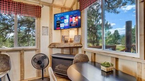A cozy cabin interior with a fireplace, a television mounted on the wall, a dining table, and large windows offering a view of trees. - Heavenly Haven Treehouse (Larkspur)