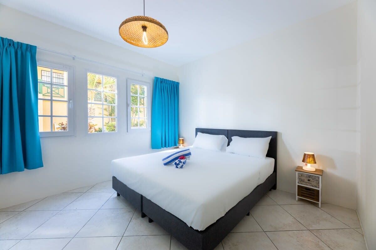 A hotel room with a large bed, two windows with blue curtains, a bedside lamp, and a hanging light fixture.