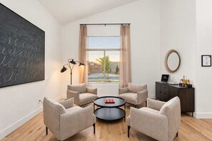 Uma sala de estar moderna com uma janela grande, uma mesa de centro redonda, duas poltronas, um abajur alto escuro e um armário de madeira escura com objetos decorativos.