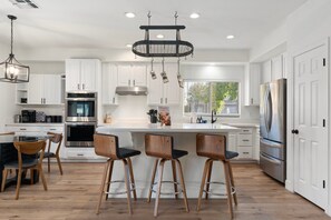 Una cocina moderna con gabinetes blancos, electrodomésticos de acero inoxidable, una isla central con taburetes y una lámpara colgante.