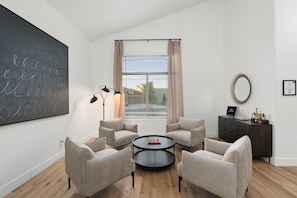 Una sala de estar moderna con una pizarra grande, una mesa de centro redonda, cuatro sillones, una lámpara de pie, un ventanal con persianas, un aparador de madera oscura y un espejo en la pared.