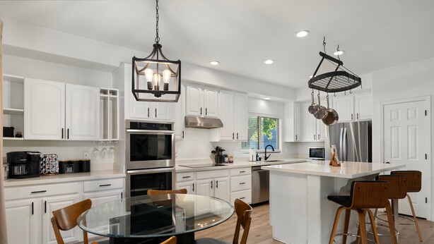 A modern kitchen with white cabinetry, stainless steel appliances, a glass dining table, and two bar stools. - The Cardinal | Ultimate Group Retreat in Glendale (Glendale)