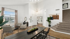 A spacious living room with a dark wooden dining table, comfortable seating, a large window with curtains, and a staircase leading to another level. - The Cardinal | Ultimate Group Retreat in Glendale (Glendale)