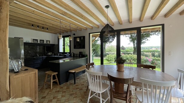 A modern kitchen with wooden beams, a large island, and a dining area with white chairs and a table. - Renovated country property with fireplace, near forest (Le Mont-Saint-Adrien)