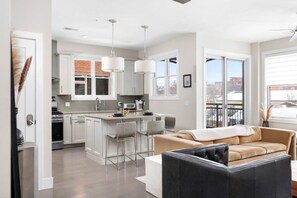 A modern kitchen with white cabinets, a central island with bar stools, pendant lighting, and a view of a cityscape through large windows. - Park 202  Downtown in the Heart of Traverse City (Traverse City)