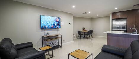 A modern living room with a flat-screen TV, black leather sofas, a coffee table, and a dining area with a small table and chairs.