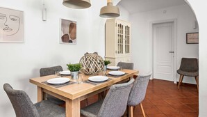 A dining area with a wooden table set for four, grey upholstered chairs, two pendant lights, a cabinet, and framed artwork on the wall.