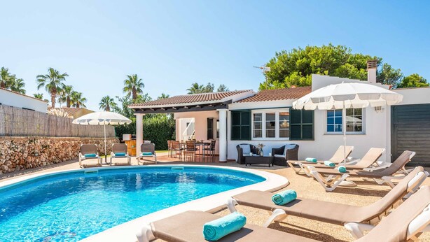 A swimming pool with lounge chairs and umbrellas, a patio area with outdoor furniture, and a building with a tiled roof.