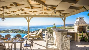 An outdoor patio with a view of the sea, a stone barbecue, and a dining area with tables and chairs.