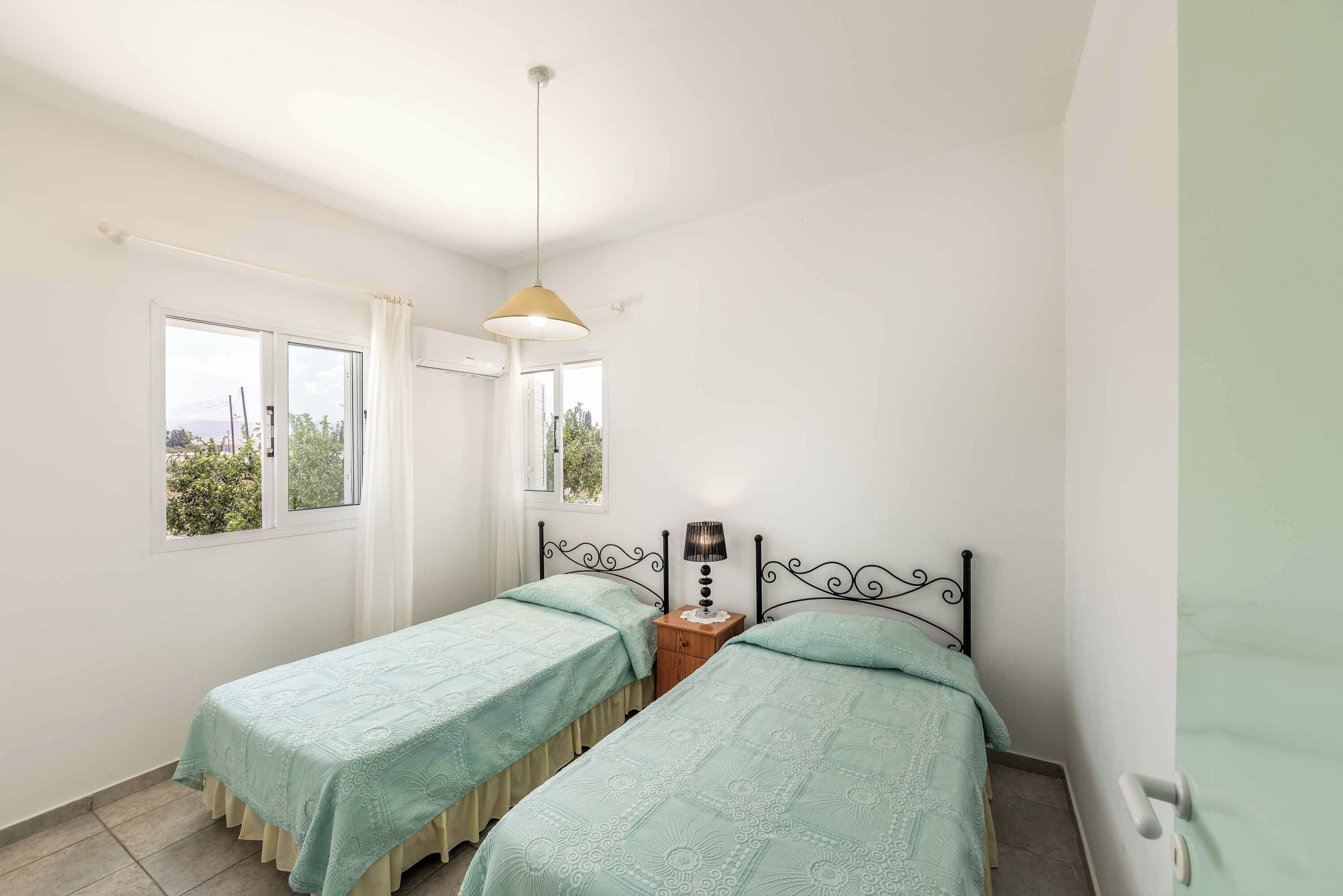 A bedroom with two single beds, a bedside table with a lamp, a window with curtains, and a hanging light fixture.