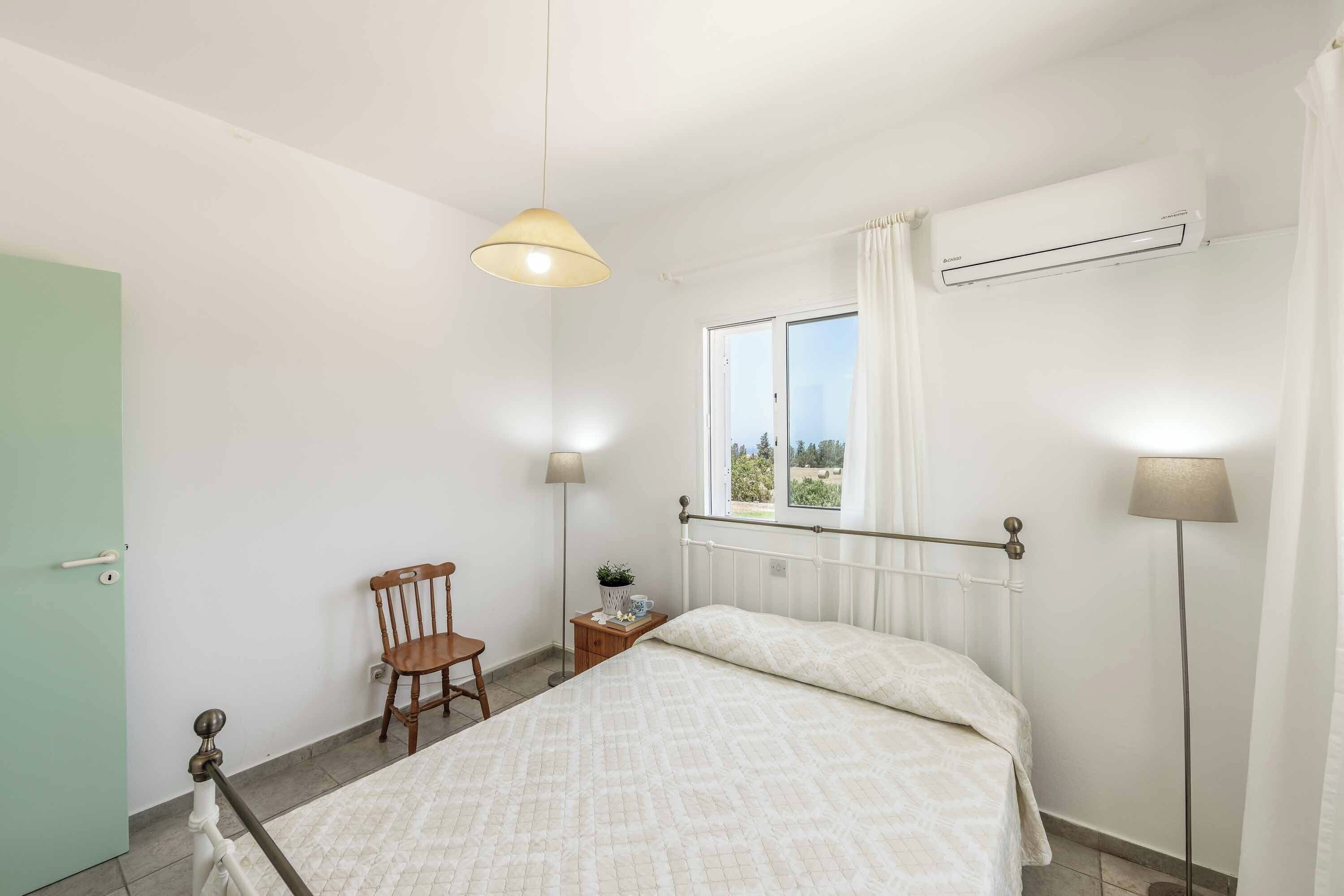 A bedroom with a bed, a chair, a bedside table, a lamp, and an air conditioning unit.