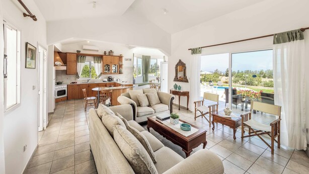 Un salón espacioso con sofá, mesa de centro y comedor. Se ve una cocina al fondo con vistas al exterior.