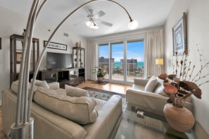 A modern living room with a large TV, comfortable sofas, a glass coffee table, and a view of the ocean.
