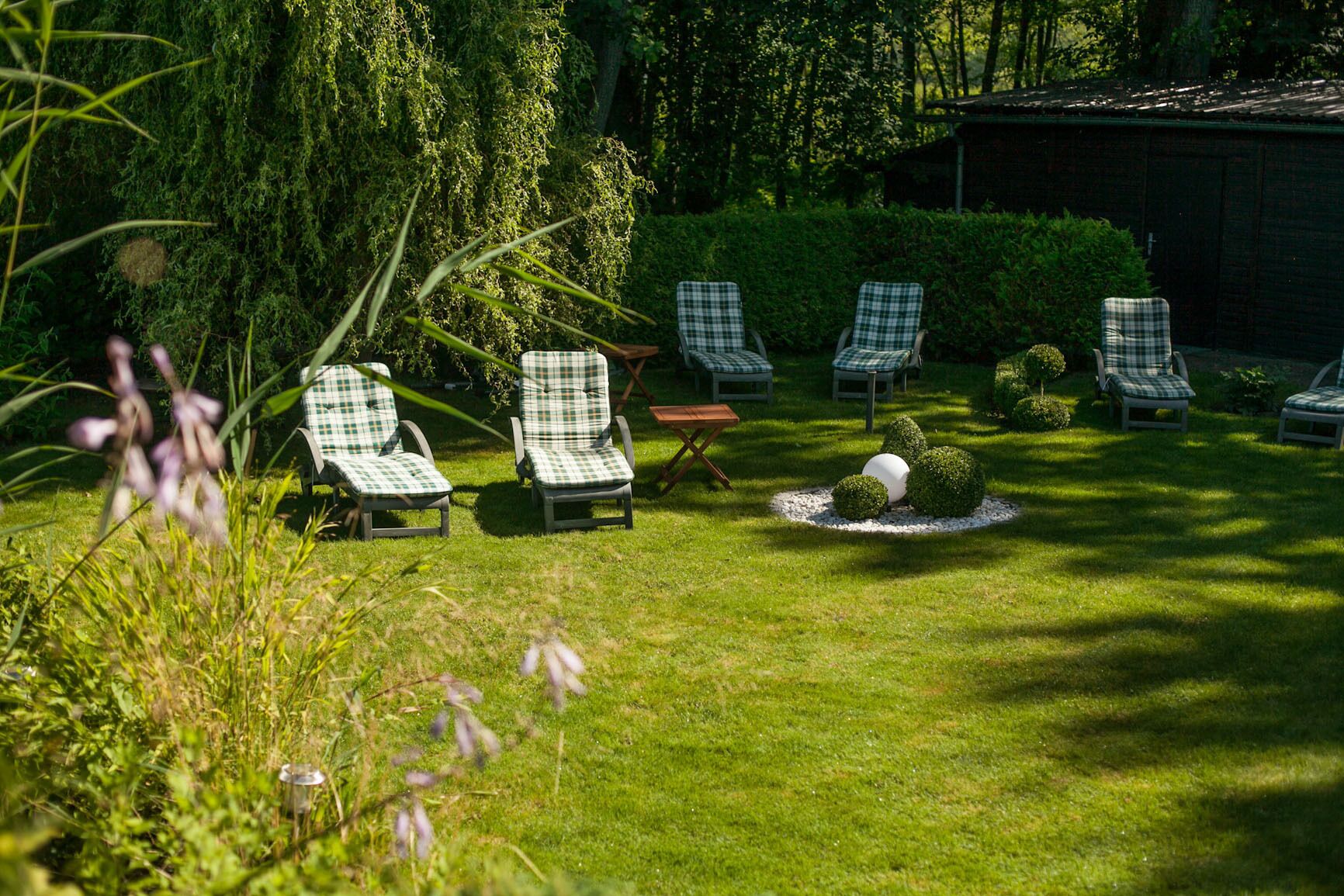 A garden with several checkered lounge chairs arranged around a central table, surrounded by well-maintained hedges and a small spherical sculpture.