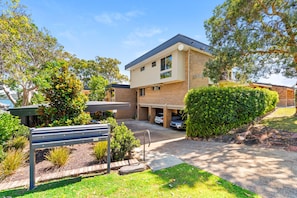 A modern two-story building with a flat roof, a covered parking area, and a well-maintained garden with a bench and various shrubs.