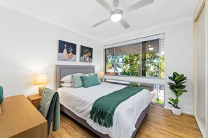 Een moderne slaapkamer met een bed, nachtkastjes, een plafondventilator en een groot raam met uitzicht op groen.