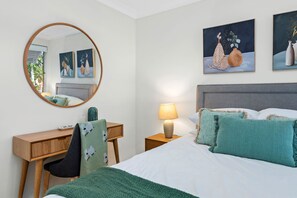 Ein Schlafzimmer mit einem Bett, einem Schreibtisch, einem Stuhl, einem Nachttisch mit Lampe, einem runden Spiegel und zwei an der Wand befestigten Bildern.