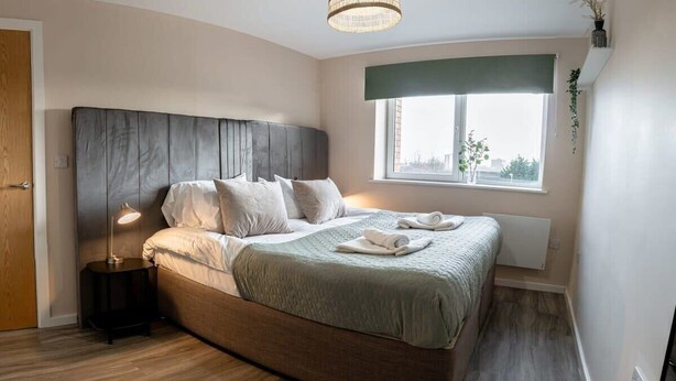 A modern bedroom with a large bed, a bedside lamp, a window with a view, and a wooden floor. - Modern & Spacious 1 Bed Apartment in Manchester (Salford)
